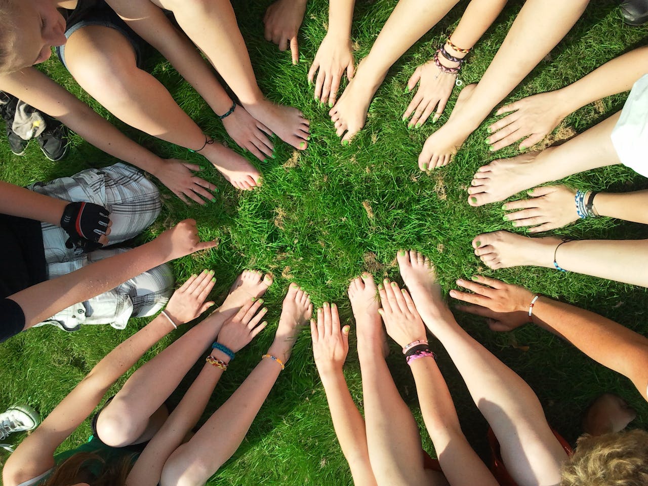 about-01 Group of friends forming a circle with their hands and feet on grass, symbolizing unity and friendship.